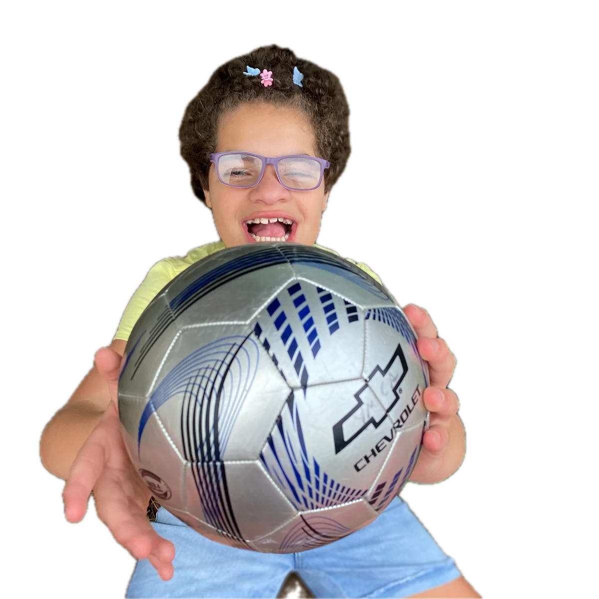 A smiling young girl in glasses holds out a silver soccer ball toward the camera in a sunlit yard.